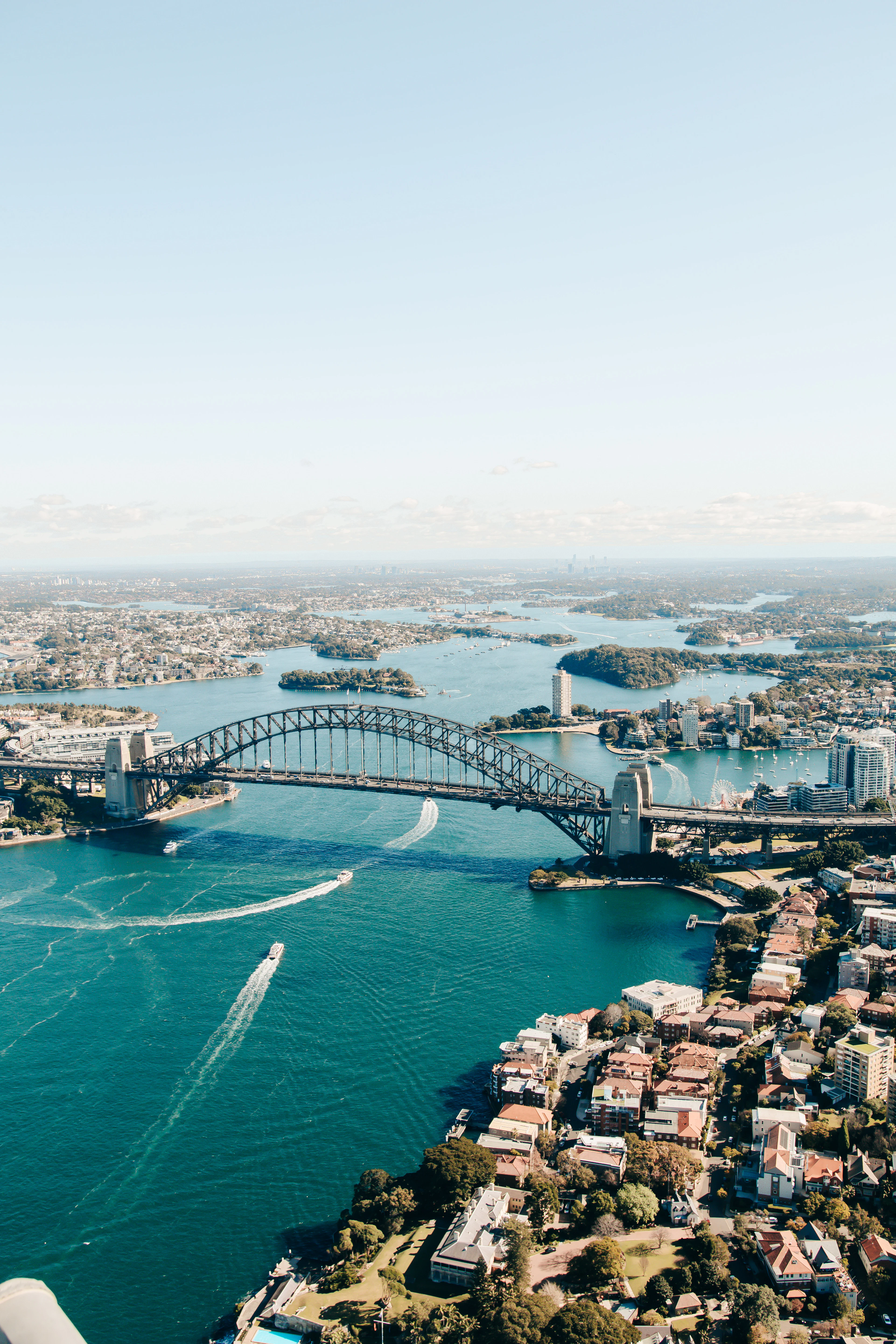 Aerial view of Sydney Harbour Bridge with boats crossing the turquoise water and harbourside suburbs beyond.