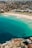 A picturesque coastal cityscape featuring a beach with golden sand and vibrant turquoise waters. The shoreline is lined with buildings, transitioning into an urban area filled with dense residential and commercial structures. The ocean has varying shades of blue, with some white waves near the shore. Above, the city stretches into the distance with numerous high-rises and greenery.