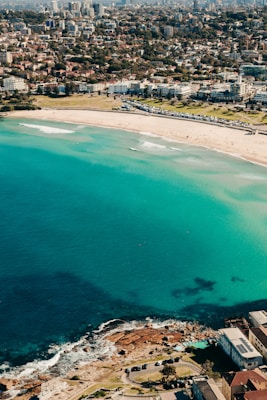 A picturesque coastal cityscape featuring a beach with golden sand and vibrant turquoise waters. The shoreline is lined with buildings, transitioning into an urban area filled with dense residential and commercial structures. The ocean has varying shades of blue, with some white waves near the shore. Above, the city stretches into the distance with numerous high-rises and greenery.