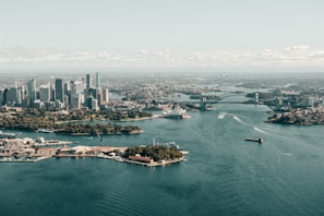 Aerial view of a large city featuring a harbor with iconic architectural landmarks, modern skyscrapers, and expansive water. Boats are visible navigating the harbor, and the landscape includes urban and green spaces.