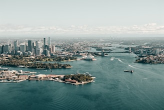 Aerial view of a large city featuring a harbor with iconic architectural landmarks, modern skyscrapers, and expansive water. Boats are visible navigating the harbor, and the landscape includes urban and green spaces.
