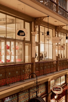 An indoor shopping arcade with intricate iron railings and vintage hanging lanterns. The scene features store signs including 'Dinosaur Designs' and 'The Nut Shop'. The upper floor displays apparel and decorative items in the shop windows, while stained glass accents adorn the lower level.