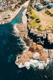 An aerial view captures a coastal area with a rocky shoreline meeting a vibrant blue ocean. Residential buildings are visible on the right, surrounded by roads and vegetation. Waves crash against the shoreline, creating white foam, while a small beachfront or harbor area is present.