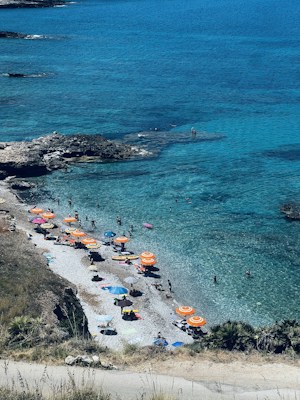 A beautifully clear and tranquil beach scene with turquoise waters and rocky shorelines. The beach is lined with vibrant orange and blue beach umbrellas, providing shade for people relaxing on the pebbly sand. In the foreground, a small path leads down to the beach, surrounded by dry grass and sparse shrubs.