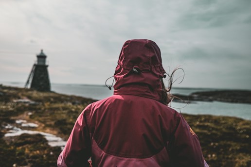 A person wearing a maroon jacket with a hood up stands on a rocky landscape near a body of water. A lighthouse is visible in the distance on the left, while the sky is overcast, giving a moody ambiance.