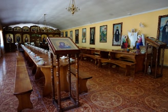 A long hall features wooden tables and benches, arranged in a dining setup with multiple tea cups and saucers. The walls are adorned with framed religious icons and paintings. At one end, religious statues and a stand holding an image are prominent. Chandeliers hang from the ceiling, illuminating the room with a warm glow.