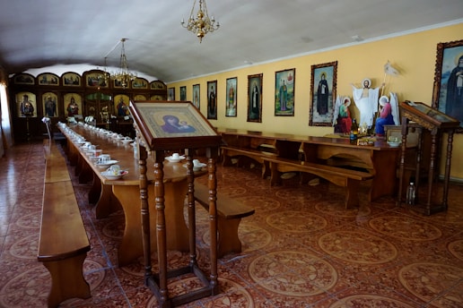 A long hall features wooden tables and benches, arranged in a dining setup with multiple tea cups and saucers. The walls are adorned with framed religious icons and paintings. At one end, religious statues and a stand holding an image are prominent. Chandeliers hang from the ceiling, illuminating the room with a warm glow.