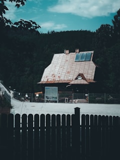A rustic cabin with a steep, metal roof stands amidst dense greenery. The house has large windows and a few solar panels on the roof. A gravel pathway leads to the cabin, with a fence in the foreground. There are informational boards and a few people in the background, suggesting a tourist or park area.