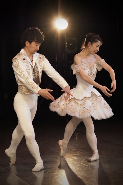 A male and female ballet dancer are performing on stage under a bright spotlight. The male dancer is dressed in a white costume with golden embroidery, while the female dancer is wearing a white tutu with matching embroidery. Both dancers are captured in a graceful pose with their arms elegantly positioned.