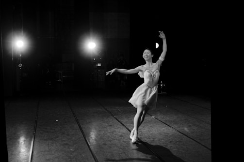 A ballet dancer gracefully poses on stage, captured in black and white. The spotlight on the dancer creates a dramatic contrast against the darkened background. The dancer's expression is serene and focused, with arms elegantly extended and one foot slightly off the floor. The composition highlights the beauty and poise of the performance.