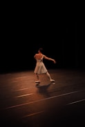 A ballet dancer is performing on a dimly lit stage, wearing a white dress and ballet shoes. The dancer's arms are extended gracefully, and the stage is mostly dark, highlighting the dancer as they move.