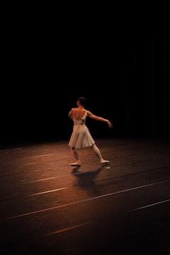 A ballet dancer is performing on a dimly lit stage, wearing a white dress and ballet shoes. The dancer's arms are extended gracefully, and the stage is mostly dark, highlighting the dancer as they move.