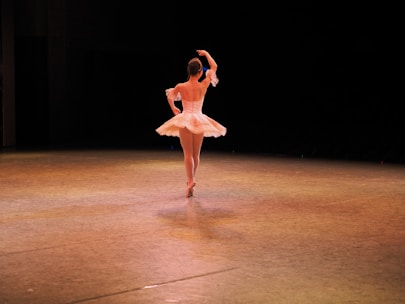 A ballet dancer is performing on stage, wearing a white tutu and pointe shoes. The scene is illuminated by warm stage lights, highlighting the dancer's graceful pose against a dark background.