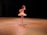 A ballet dancer is performing on stage, wearing a white tutu and pointe shoes. The scene is illuminated by warm stage lights, highlighting the dancer's graceful pose against a dark background.
