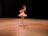 A ballet dancer is performing on stage, wearing a white tutu and pointe shoes. The scene is illuminated by warm stage lights, highlighting the dancer's graceful pose against a dark background.