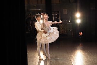 A ballet dancer in a white and gold tutu is gracefully poised alongside a male dancer in similar attire. They are in a dimly lit backstage area with stage lights glowing in the background, creating a dramatic and artistic atmosphere.