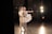 A ballet dancer in a white and gold tutu is gracefully poised alongside a male dancer in similar attire. They are in a dimly lit backstage area with stage lights glowing in the background, creating a dramatic and artistic atmosphere.