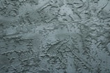 A textured surface with an irregular pattern of raised and recessed areas, resembling stucco or plaster. The surface has a rough, uneven texture with shadows enhancing the depth of the pattern.