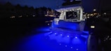 A yacht is illuminated with bright blue lights, casting a vibrant glow on the water at night. The surrounding area appears dimly lit with the silhouettes of nearby houses visible in the background.
