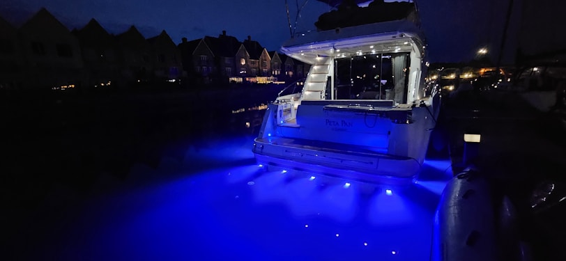 A yacht is illuminated with bright blue lights, casting a vibrant glow on the water at night. The surrounding area appears dimly lit with the silhouettes of nearby houses visible in the background.