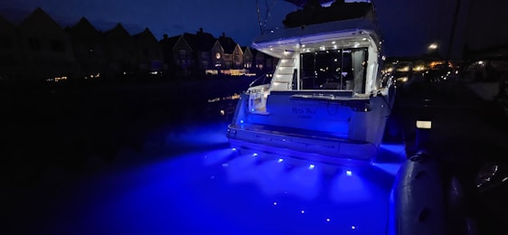 A yacht is illuminated with bright blue lights, casting a vibrant glow on the water at night. The surrounding area appears dimly lit with the silhouettes of nearby houses visible in the background.