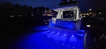 A yacht is illuminated with bright blue lights, casting a vibrant glow on the water at night. The surrounding area appears dimly lit with the silhouettes of nearby houses visible in the background.