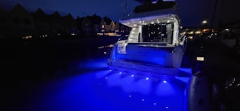 A yacht is illuminated with bright blue lights, casting a vibrant glow on the water at night. The surrounding area appears dimly lit with the silhouettes of nearby houses visible in the background.