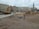 A construction site in an urban area with a large excavator, piles of dirt, pipes, and construction materials scattered around. Several people can be seen in the background near the buildings. The background includes multi-story residential and commercial buildings, and a tall monument with a statue atop it.