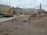 A construction site in an urban area with a large excavator, piles of dirt, pipes, and construction materials scattered around. Several people can be seen in the background near the buildings. The background includes multi-story residential and commercial buildings, and a tall monument with a statue atop it.