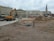 A construction site in an urban area with a large excavator, piles of dirt, pipes, and construction materials scattered around. Several people can be seen in the background near the buildings. The background includes multi-story residential and commercial buildings, and a tall monument with a statue atop it.