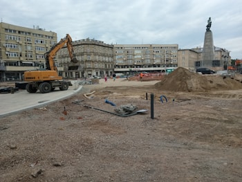 A construction site in an urban area with a large excavator, piles of dirt, pipes, and construction materials scattered around. Several people can be seen in the background near the buildings. The background includes multi-story residential and commercial buildings, and a tall monument with a statue atop it.