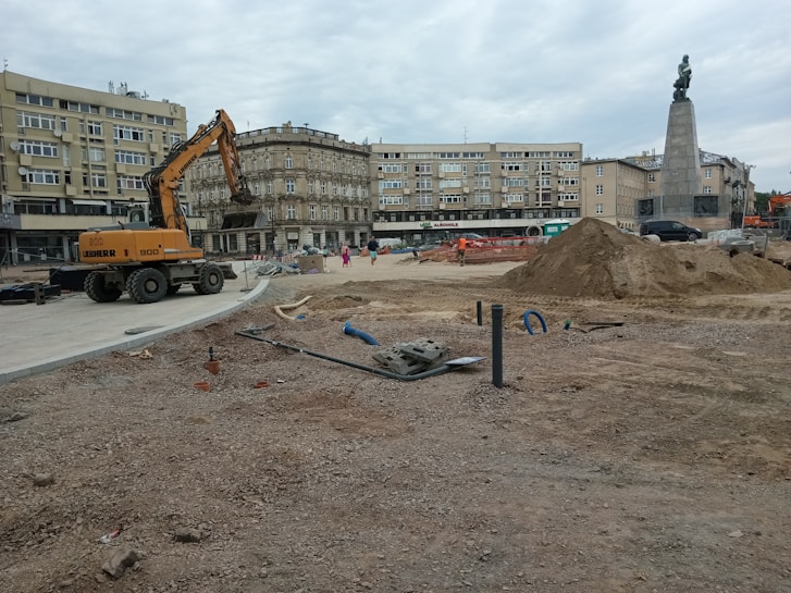A construction site in an urban area with a large excavator, piles of dirt, pipes, and construction materials scattered around. Several people can be seen in the background near the buildings. The background includes multi-story residential and commercial buildings, and a tall monument with a statue atop it.