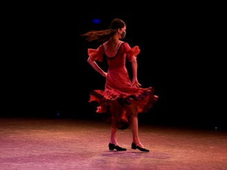 A dancer wearing a vibrant red dress twirls gracefully on stage. Her hair flows behind her as she spins, creating a sense of dynamic movement. The dark background makes the dancer stand out, highlighting the fluid motion and elegance of her performance.