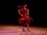 A dancer wearing a vibrant red dress twirls gracefully on stage. Her hair flows behind her as she spins, creating a sense of dynamic movement. The dark background makes the dancer stand out, highlighting the fluid motion and elegance of her performance.