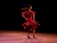 A dancer wearing a vibrant red dress twirls gracefully on stage. Her hair flows behind her as she spins, creating a sense of dynamic movement. The dark background makes the dancer stand out, highlighting the fluid motion and elegance of her performance.