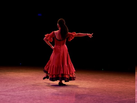 A dancer is captured performing on stage, wearing a flowing red dress with ruffles. The lighting creates a dramatic atmosphere, highlighting the movement and elegance of the dancer. The background is dark, emphasizing the focus on the performer.