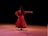 A dancer is captured performing on stage, wearing a flowing red dress with ruffles. The lighting creates a dramatic atmosphere, highlighting the movement and elegance of the dancer. The background is dark, emphasizing the focus on the performer.