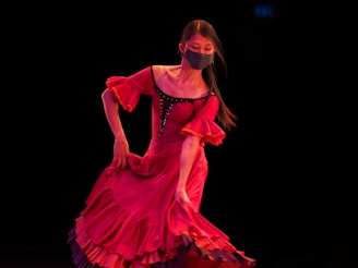 A woman is dancing gracefully while wearing a vibrant red dress with ruffles and a dark face mask. Her long hair flows as she moves against a dark background, creating a striking contrast.