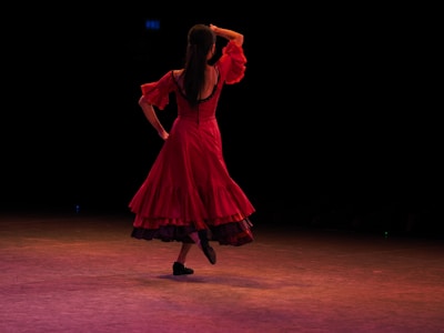 A person in a vibrant red dress is dancing on a stage with a dark background. The dancer is captured in a dynamic pose with one hand resting on their head and the other on their hip. The stage lighting casts warm tones across the floor.