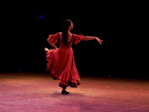 A dancer wearing a flowing red dress performs gracefully on stage, illuminated by stage lighting. The background is dark, emphasizing her elegant movements and the vibrancy of her dress.