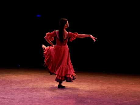 A dancer wearing a flowing red dress performs gracefully on stage, illuminated by stage lighting. The background is dark, emphasizing her elegant movements and the vibrancy of her dress.