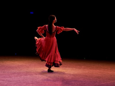 A dancer wearing a flowing red dress performs gracefully on stage, illuminated by stage lighting. The background is dark, emphasizing her elegant movements and the vibrancy of her dress.