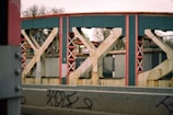 A close-up view of a steel bridge structure with large metal beams and rivets, featuring a mix of red and teal-colored components. The foreground shows a concrete barrier with graffiti, and in the background, bare trees and an overcast sky can be seen.