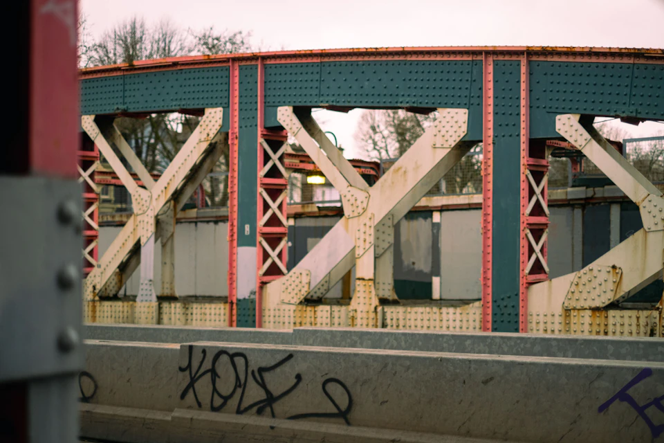 The image shows a bridge with a unique design, featuring red and white stripes on its sides and green railings. The bridge is supported by metal beams that are rusted, indicating it has been exposed to the elements for some time. In front of the bridge, there's graffiti covering the concrete wall, adding an urban touch to the scene.

The perspective of the image is from a train track, looking towards the bridge. This vantage point provides a clear view of both the bridge and its surroundings, showcasing the contrast between the industrial metal beams and the vibrant colors of the red and white stripes on the bridge's sides.