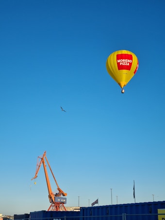 A yellow hot air balloon advertising 'Moreno Pizza' floats in a clear blue sky. Below it, an industrial scene with a large orange crane is visible. A bird can be seen flying in the sky near the balloon. The bottom of the image shows blue industrial buildings or containers.