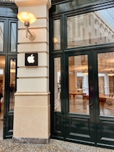 A modern retail storefront with a black-framed glass facade featuring a prominent Apple logo mounted on a cream-colored wall. The interior shows wooden tables with electronic devices and a spacious, well-lit environment.