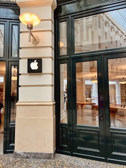 A modern retail storefront with a black-framed glass facade featuring a prominent Apple logo mounted on a cream-colored wall. The interior shows wooden tables with electronic devices and a spacious, well-lit environment.
