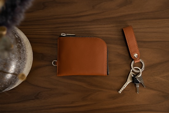 A brown leather wallet is placed on a dark wooden surface next to a matching leather keychain with three metal keys attached. A decorative vase or object partially appears on the left edge, adding a touch of elegance to the setting.
