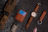 Various everyday carry items are neatly arranged on a dark, textured surface. A large knife with a brown handle and sheath, a brown leather wallet holding a blue credit card, and a brown leather wristwatch with a silver face are visible. A multi-tool, a brown leather keychain, and a red Swiss Army knife are also among the items.
