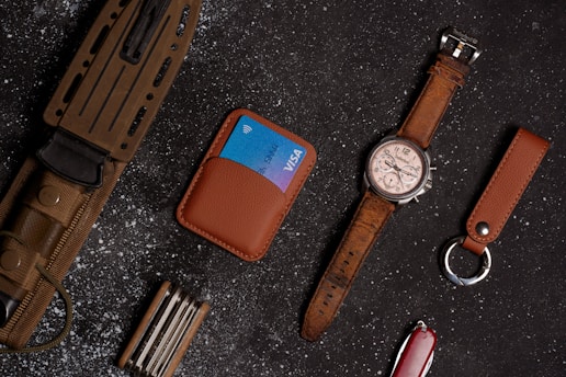 Various everyday carry items are neatly arranged on a dark, textured surface. A large knife with a brown handle and sheath, a brown leather wallet holding a blue credit card, and a brown leather wristwatch with a silver face are visible. A multi-tool, a brown leather keychain, and a red Swiss Army knife are also among the items.